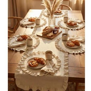Könnte beinhalten: Ein hölzerner Esstisch mit einem beigen Leinentischläufer und passenden runden Platzsets mit gerüschten Rändern. Jedes Gedeck besteht aus einem Keramikteller mit einem Croissant und Gebäck, einer Keramiktasse und einer gefalteten Leinenserviette.
