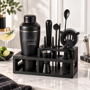 Op de afbeelding: Een matzwarte cocktailshaker-set met een shaker voorzien van de gegraveerde tekst Michael, 19.08.2026, GROOMSMAN, een jigger, stamper, barlepel, zeef en kurkentrekker, alles georganiseerd in een zwarte houten standaard op een marmeren ondergrond.
