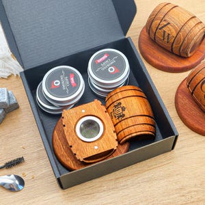 May include: A black gift box containing two round metal tins of smoking chips, a wooden cigar cutter, and a wooden barrel-shaped cup. Outside the box are two additional wooden barrel-shaped cups on coasters, three grey stone cubes, and a small brush.
