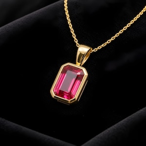 May include: A gold pendant necklace featuring a rectangular, emerald-cut vibrant pink gemstone set in a polished gold bezel. The pendant hangs from a delicate gold chain, showcasing a clean, geometric design with a bright, reflective finish.