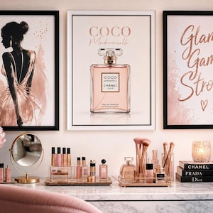 Three framed wall art prints featuring a silhouetted woman, a perfume bottle, and pink script text hanging above a makeup vanity table.