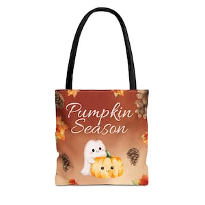 Könnte beinhalten: Eine Tragetasche mit schwarzen Henkeln und braunem Hintergrund, auf dem der Text Pumpkin Season in weißer Schreibschrift steht. Das Motiv zeigt ein kleines weißes Gespenst, das einen orangefarbenen Kürbis umarmt, umgeben von Herbstblättern und Tannenzapfen.