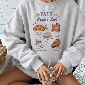 Könnte beinhalten: Ein hellgraues Sweatshirt mit Rundhalsausschnitt und einem Aufdruck mit der Aufschrift FALL Bucket List. Das Design zeigt Illustrationen von einem Laubhaufen, einem Korb mit Äpfeln, einem Kuchen, einer Tasse Apfelwein und Kürbissen mit Text.
