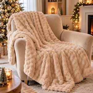 Può includere: Una morbida coperta in pelliccia sintetica color crema con un motivo a coste testurizzato, drappeggiata su una poltrona imbottita abbinata. La coperta presenta un pelo folto e soffice che copre la seduta e lo schienale della poltrona in modo stratificato.