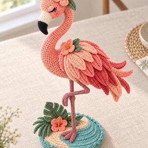 May include: A crochet flamingo figurine featuring a pink body, a black and white beak, and closed eyes with pink cheeks. The flamingo is adorned with pink hibiscus flowers and green leaves, standing on a circular base with blue and beige yarn.