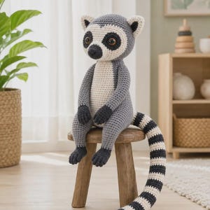 May include: A crocheted lemur stuffed animal featuring a grey and white body, dark grey hands and feet, and large brown eyes. The lemur has a long, prominent tail with alternating black and white stripes, sitting upright on a small wooden stool.