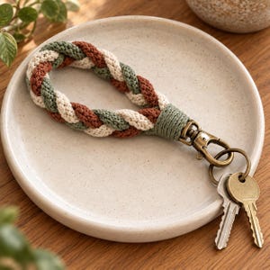 May include: A braided keychain featuring three-tone cord in sage green, terracotta, and cream. The loop is secured with a sage green wrapped knot and attached to an antique brass swivel lobster clasp holding two metal keys.