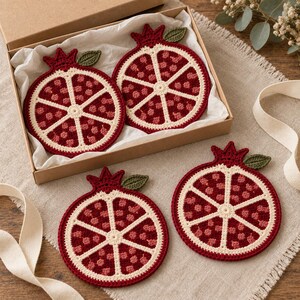 May include: Four round, crocheted pomegranate coasters featuring a cross-section design with deep red and light pink seeds, a cream-colored rind, and a small green leaf attached to the top. Two coasters are inside a box and two are outside.