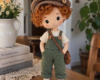 May include: A handcrafted crochet doll with curly ginger hair, wearing a brown newsboy cap, a cream-colored shirt, and olive green overalls. The doll features a small brown crossbody bag, brown shoes, and has rosy cheeks with black button eyes.