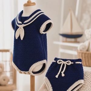May include: A dark blue crochet sailor-style bodysuit with a white collar, short sleeves, and a matching pair of dark blue crochet shorts with a white drawstring waist. The bodysuit features three small buttons at the bottom leg opening.