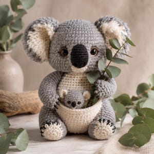 Pode incluir: Um brinquedo de koala em crochê nas cores cinza e creme, apresentando um koala adulto com uma bolsa contendo um pequeno koala bebé. O koala adulto tem orelhas grandes e felpudas, olhos escuros e um nariz preto, segurando um ramo de eucalipto.