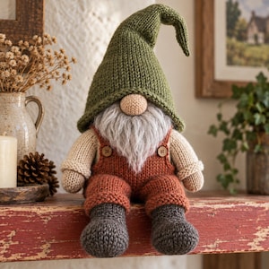 Puede incluir: Un muñeco gnomo tejido con un gorro puntiagudo de color verde oliva, cara beige con nariz redonda y una larga barba gris. El gnomo viste un peto de color óxido con dos pequeños botones marrones sobre una camisa beige de manga larga y botas gris oscuro.