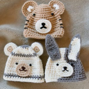 May include: Three crocheted hats featuring animal designs. The top hat is tan with bear ears and a white muzzle. The bottom left hat is white with gray stripes and bear ears. The bottom right hat is white and gray with long rabbit ears.