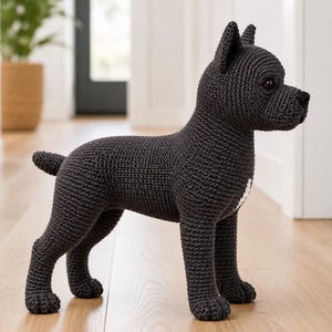 Könnte beinhalten: Eine dunkel anthrazitfarbene Häkelfigur eines Hundes im Profil. Das handgefertigte Stofftier hat eine texturierte Garnoberfläche, spitze Ohren, einen kurzen Schwanz und einen kleinen weißen Fleck aus Garn auf der Brust.