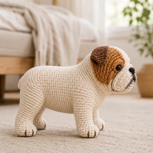 May include: A crochet bulldog figurine featuring a cream-colored body with brown patches on the head and ears. The stuffed animal has black bead eyes, a black stitched nose, and a short tail, crafted with a visible yarn texture throughout.
