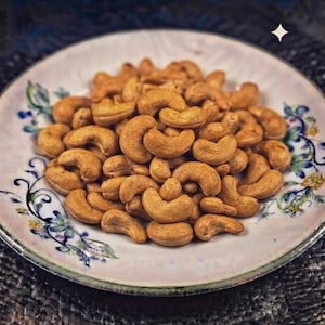 Könnte beinhalten: Ein Haufen gerösteter, goldbrauner Cashewkerne in der Mitte eines runden, weißen Keramiktellers. Der Teller ist mit einem zarten Blumenmuster aus blauen und grünen Blättern sowie kleinen gelben Blüten am geschwungenen Rand verziert.