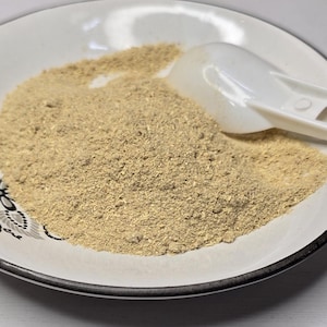 May include: A white ceramic bowl with a black rim and a decorative pattern contains a mound of fine, light beige powder. A small, white plastic scoop rests on top of the powder, partially submerged in the granular substance within the bowl.