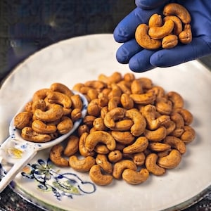 Könnte beinhalten: Ein Haufen gerösteter, goldbrauner Cashewkerne auf einem weißen Keramikteller mit Blumenmuster. Ein weißer Keramiklöffel, gefüllt mit weiteren Cashews, liegt auf dem Teller, während einige weitere Nüsse in einer blau behandschuhten Hand gehalten werden.
