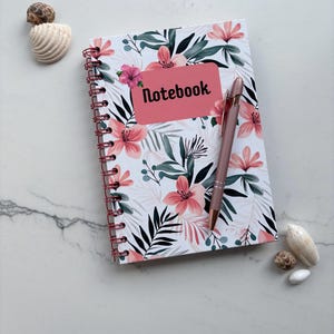 May include: A spiral-bound notebook with a white cover featuring a pink and green floral pattern, a pink rectangular label with the word Notebook printed in black, and a matching rose gold pen resting on the cover.