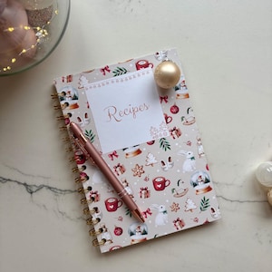 Può includere: Un taccuino a spirale con copertina beige caratterizzato da un motivo festivo di palle di neve, conigli, regali e tazze. Un'etichetta bianca al centro riporta la scritta "Recipes" in corsivo. Una penna color oro rosa poggia sulla copertina.
