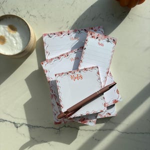 Pode incluir: Uma pilha de blocos de notas retangulares e quadrados com borda recortada e um padrão de abóboras laranja e folhas de outono. Cada bloco tem a palavra Notes impressa em letra laranja. Uma caneta esferográfica marrom repousa sobre o bloco.