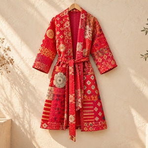 May include: A knee-length quilted robe featuring a vibrant red patchwork design with various floral and geometric patterns. The garment includes long sleeves, a matching tie belt at the waist, and two front pockets, all constructed with visible stitching.