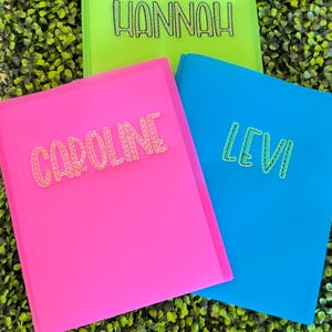 May include: Three brightly colored folders: pink, green, and blue. Each folder has a name in glittery letters: "Caroline", "Hannah", and "Levi". The folders are arranged on a bed of green foliage.