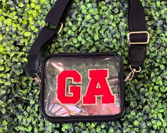 Puede incluir: Bolso transparente de plástico con correa cruzada ajustable negra. El bolso tiene un parche "GA" rojo y dorado en la parte delantera.