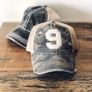 May include: Two distressed baseball caps. One is a camo pattern with a white number 9 with gold trim. The other is a black and white camo pattern.