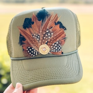 May include: A green trucker hat with a brown and black pheasant feather and a gold shotgun shell on the front.