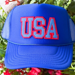 May include: A bright blue trucker hat featuring a foam front panel and mesh back panels. The front displays the text USA in large block letters with a red outline and a blue and white checkered pattern fill. A blue cord trim sits above the brim.