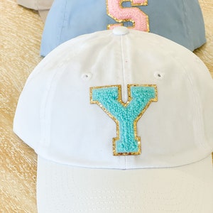 May include: Three baseball caps with different colored letters. The top cap is light blue with a pink letter "S". The middle cap is white with a turquoise letter "Y". The bottom cap is beige.