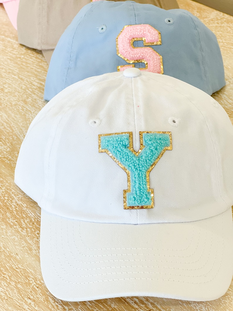 May include: Three baseball caps with different colored letters. The top cap is light blue with a pink letter "S". The middle cap is white with a turquoise letter "Y". The bottom cap is beige.