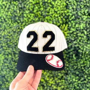 May include: Baseball cap with a black brim and white pinstripes. The cap features the number "22" in black with gold trim and a baseball patch. The background is a green hedge.