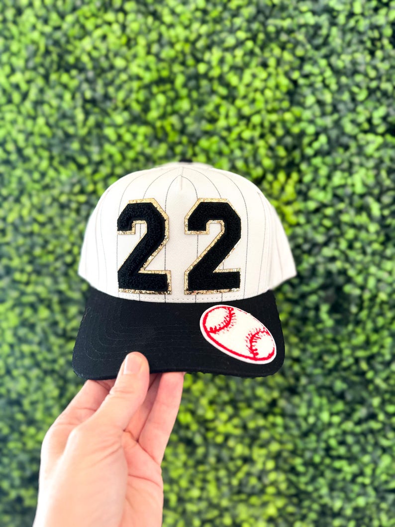 May include: Baseball cap with a black brim and white pinstripes. The cap features the number "22" in black with gold trim and a baseball patch. The background is a green hedge.
