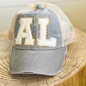 May include: A gray and beige baseball cap with a white and gold "AL" patch on the front. The cap has a mesh back and a distressed look.