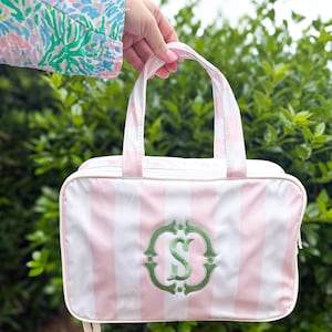 May include: A rectangular travel bag featuring a light pink and white vertical striped pattern. The front of the bag displays a centered, embroidered green monogram of the letter S within a decorative circular frame. It has two top handles.