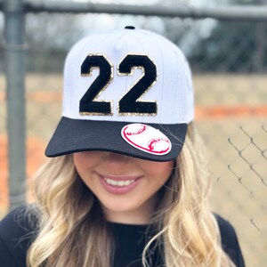 May include: White baseball cap with a black brim and the number "22" in black with gold glitter accents. A baseball sticker is on the front. The cap is worn by a person with blonde hair.