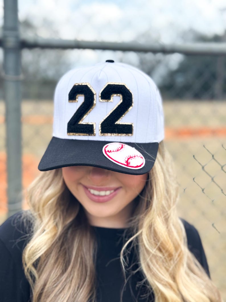 May include: White baseball cap with a black brim and the number "22" in black with gold glitter accents. A baseball sticker is on the front. The cap is worn by a person with blonde hair.