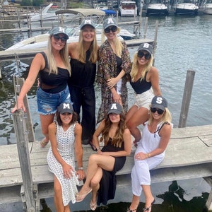 May include: A group of friends wearing gray baseball caps with white letters on a dock by a lake. The caps spell out the word "MAS"