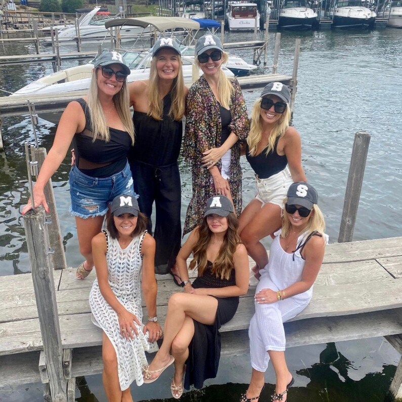 May include: A group of friends wearing gray baseball caps with white letters on a dock by a lake. The caps spell out the word "MAS"