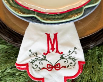 May include: A white linen napkin with a red embroidered letter "M" and decorative green and red accents. The napkin has a scalloped red border and is part of a Christmas table setting with decorative plates.