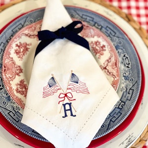 May include: A folded white linen napkin embroidered with two American flags and the letter "A" in blue and gold. The napkin is tied with a navy blue ribbon and rests on a red and white patterned plate.
