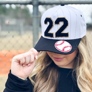 May include: A baseball cap with a white and gray pinstripe design, featuring the number "22" in black with gold glitter accents. The cap has a black brim and a baseball patch.
