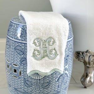 May include: A white monogrammed towel with a light green and blue embroidered initial rests on a blue and white ceramic garden stool. The stool features a detailed floral pattern.