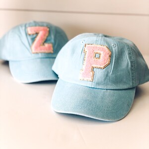 May include: Two light blue baseball caps with pink and gold letter patches. One cap has a "Z" patch, and the other has a "P" patch. The caps are made of a soft material and have a curved brim.