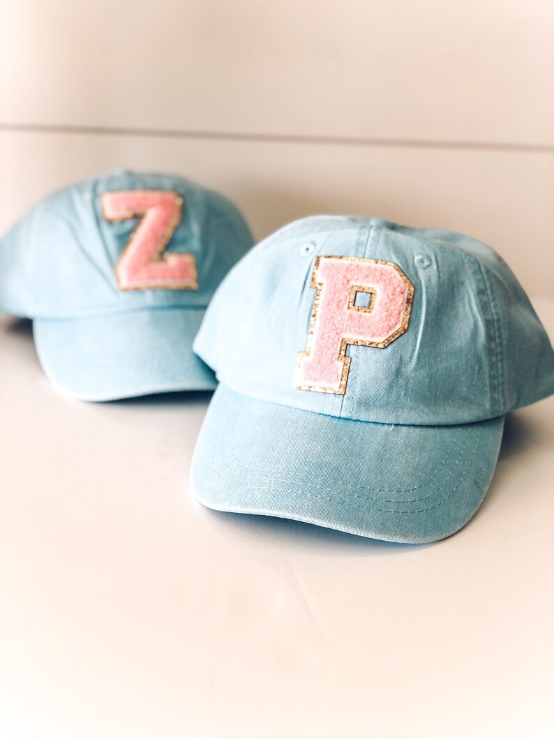 May include: Two light blue baseball caps with pink and gold letter patches. One cap has a "Z" patch, and the other has a "P" patch. The caps are made of a soft material and have a curved brim.