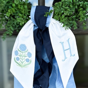 May include: A white and light blue decorative bow with a navy blue ribbon. The bow features a light blue embroidered floral design and the letter "H". The bow is hanging in front of green foliage.