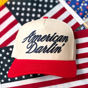 Puede incluir: Una gorra de b&eacute;isbol de color crema con un panel frontal estructurado y una visera de color rojo brillante. La parte delantera de la gorra presenta el texto "American Darlin'" bordado en una fuente cursiva azul marino oscuro.