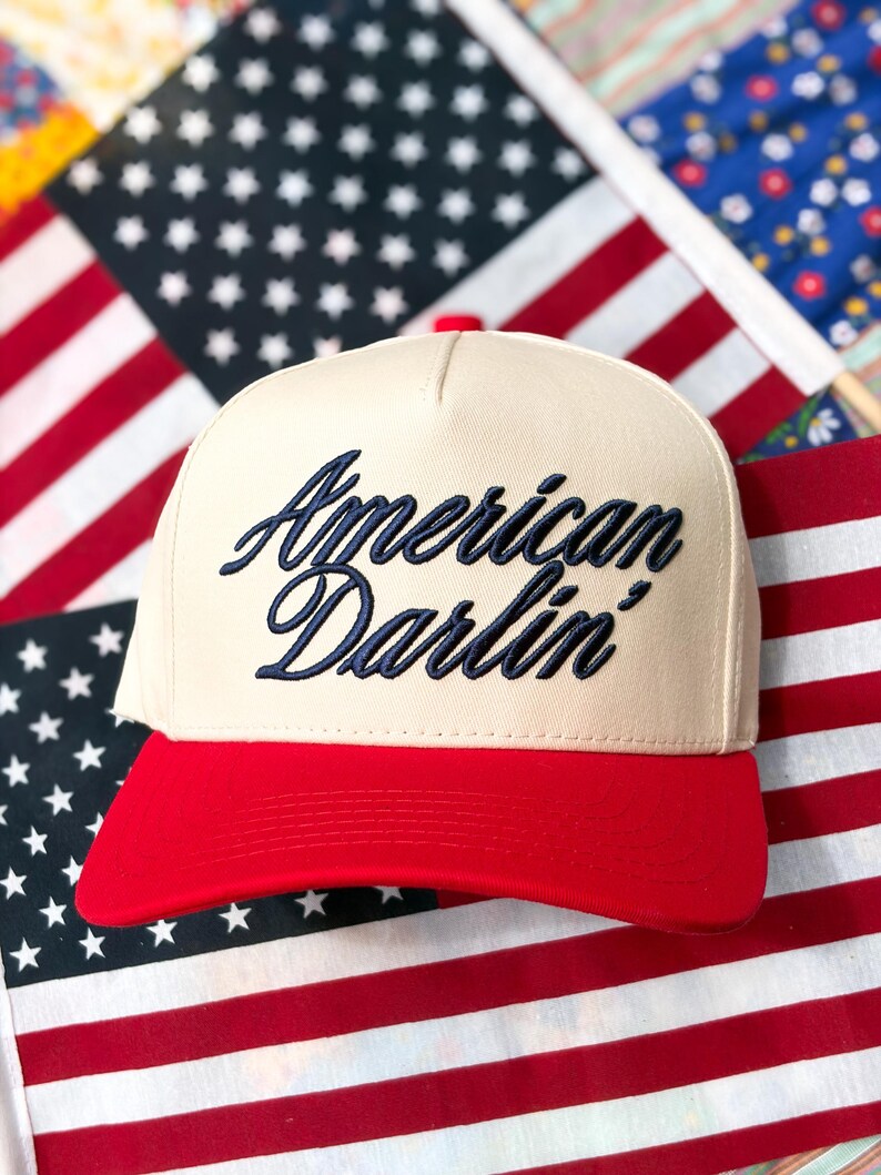 Puede incluir: Una gorra de b&eacute;isbol de color crema con un panel frontal estructurado y una visera de color rojo brillante. La parte delantera de la gorra presenta el texto "American Darlin'" bordado en una fuente cursiva azul marino oscuro.